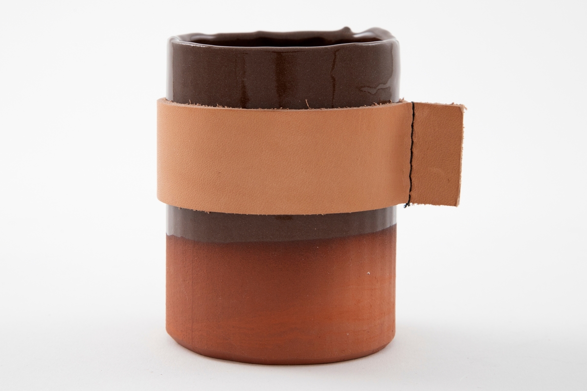 Matt brun vase i terrakotta. Sylinderform med riller. Blank brunglasert på innsiden og øvre to tredjedeler på utsiden. Midten av vasen er kledd med et bredt bånd i lysebrunt lær.
