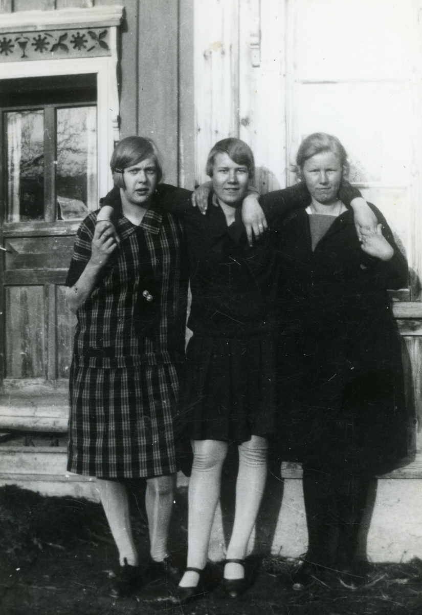 Tre unge kvinner poserar framfor ein husvegg.