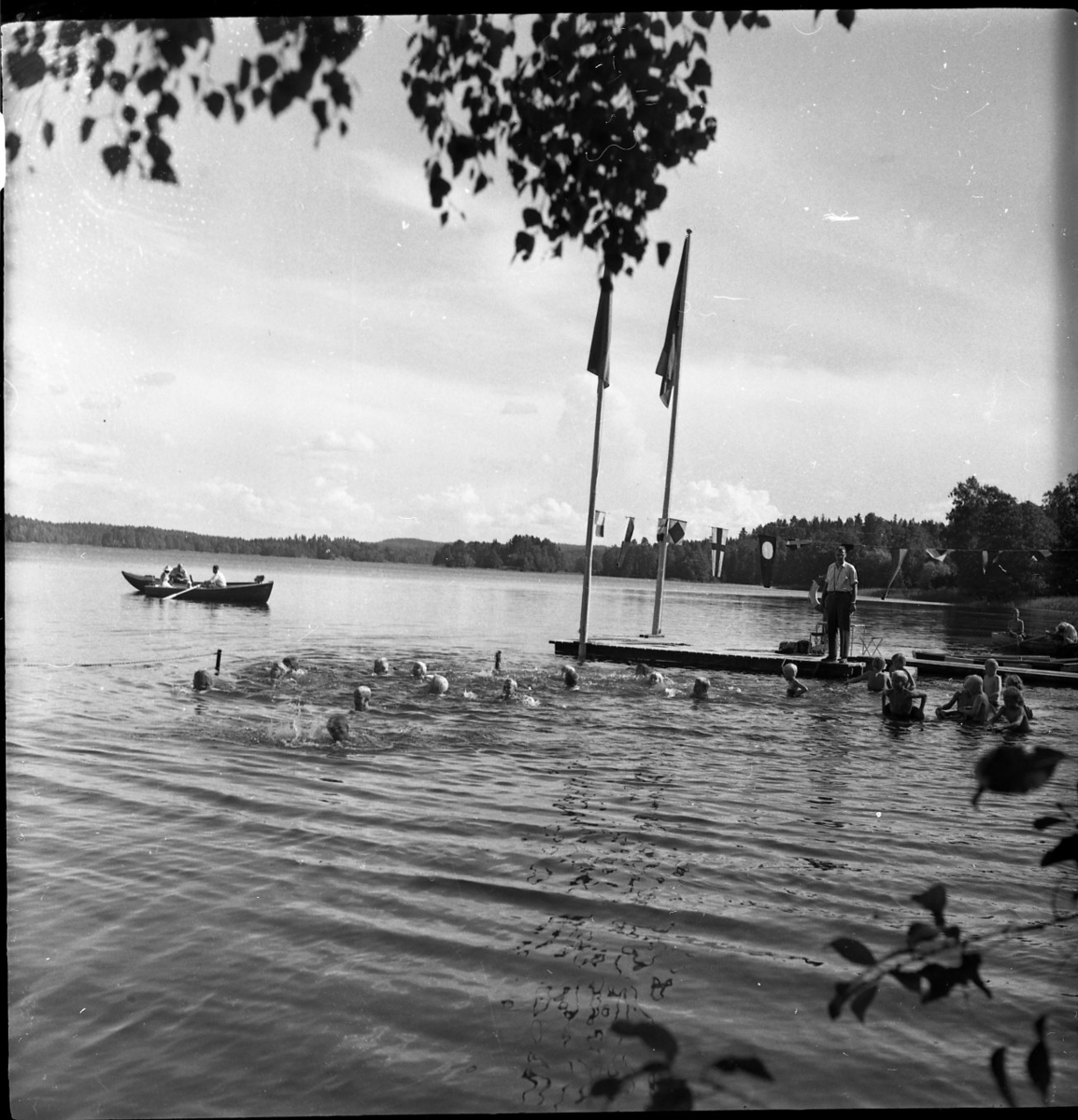 Några barn simmar i en sjö. Det står en simskollärare på en brygga och tittar på. Två flaggstänger står längst ut på bryggan. I bakgrunden syns en roddbåt.