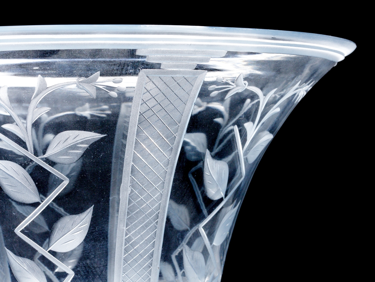 Formgiven av Edward Hald. Ljusblå trattformad skål med tillhörande fat. Skålen med slipad dekor i form av nätmönstrade pelare och stiliserade blomrankor. Nätmönstrad bård på fatets brätte.