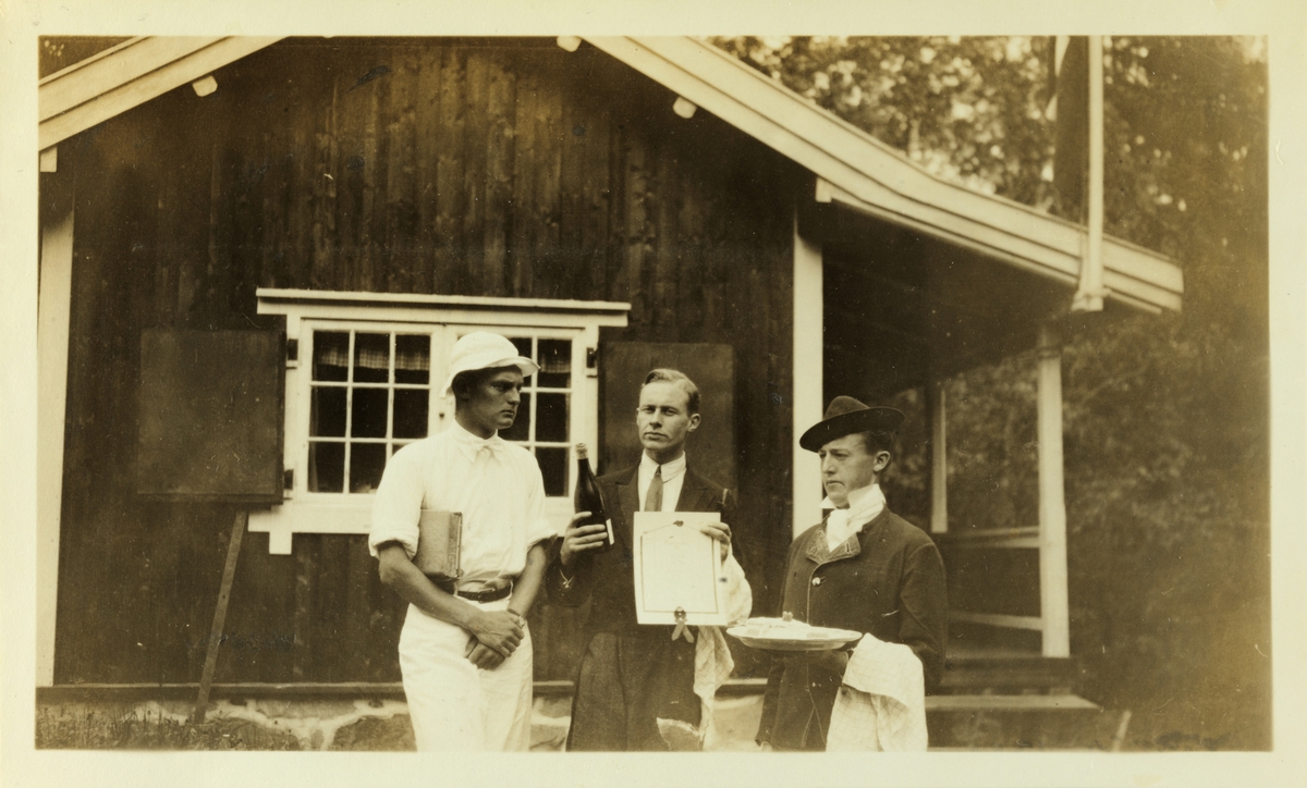 Tre unge menn, medlemmer av "Kulinarisk Klub", har stilt opp for fotografen med bl.a. vinflaske og diplom utenfor en hytte, muligens i Bærum. Fotografert mai 1926.