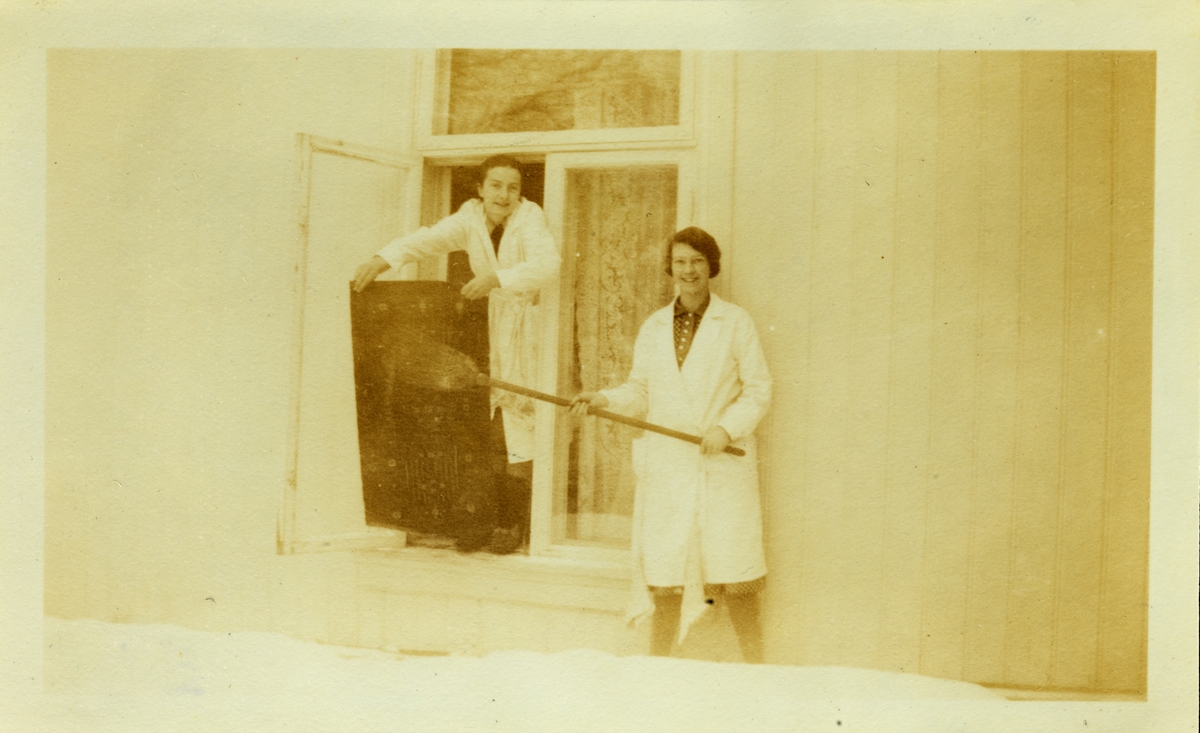 To kvinner i hvite kitler banker et teppe utenfor et vindu. Fotografert på Bjerke gård i Hedmark vinteren 1928.
