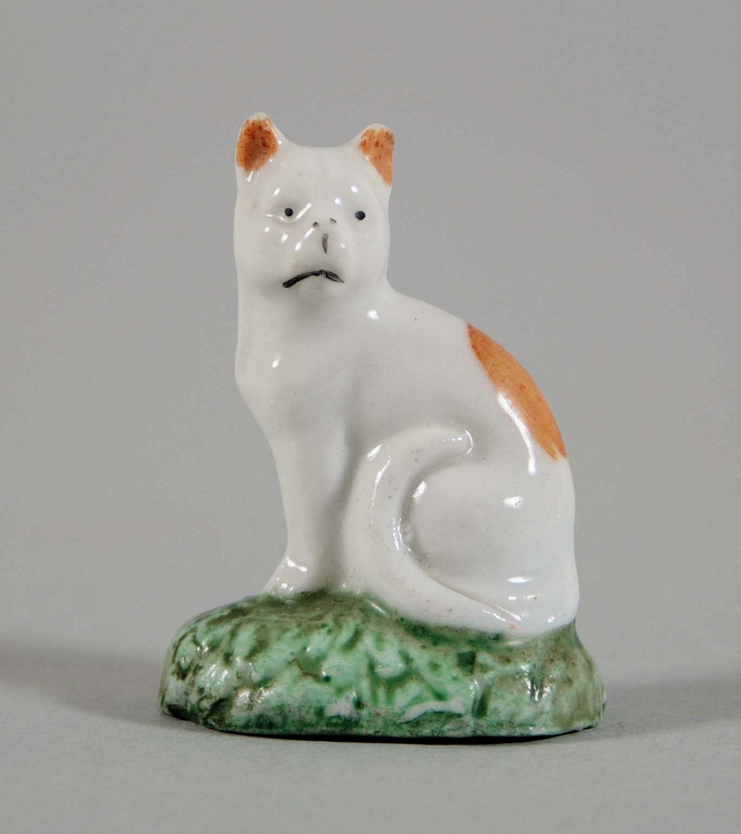 Sittande katt på oval sockel i vitglaserat porslin, med öron och ryggparti i brunt. Grönmålad sockel i form av grästuva.