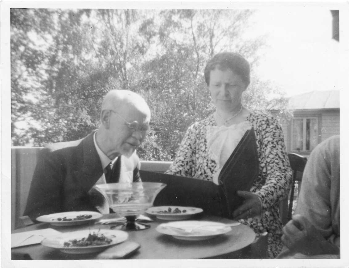 En man och en kvinna sitter på en balkong och tittar troligen i ett fotoalbum. På ett bord framför dem står det tallrikar med bär.