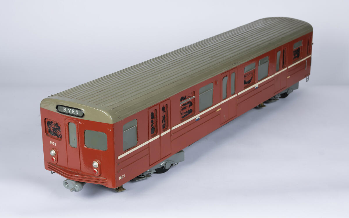Tremodell av T-banevogn nr. 1002 i en rød farge, hvit stripe langs sidene og grått tak. Under er modellen uthulet og og det er plassert to "boggier" i hver ende. 