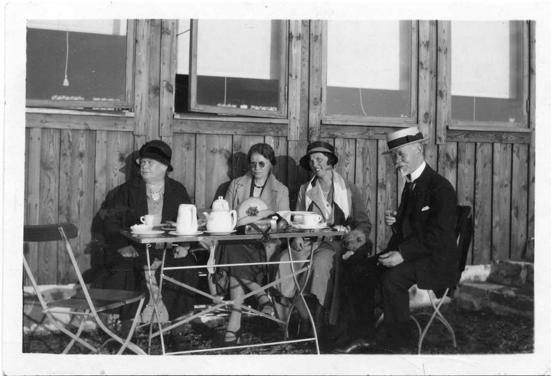Tre kvinnor och en man sitter utomhus vid ett kaffebord utmed en husvägg, i lite motljus. Kaffekanna och koppar på bordet.
