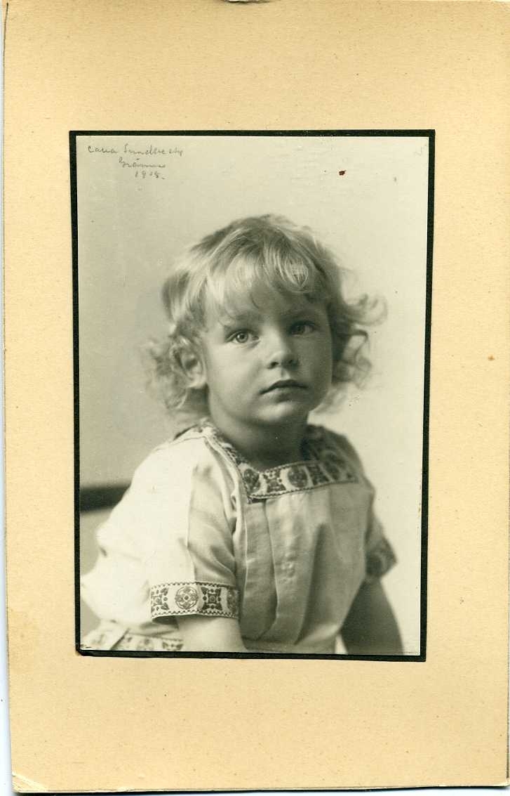 Porträtt av en ljushårig liten pojke med lockigt hår, i ljus särk med mönstrad bård.