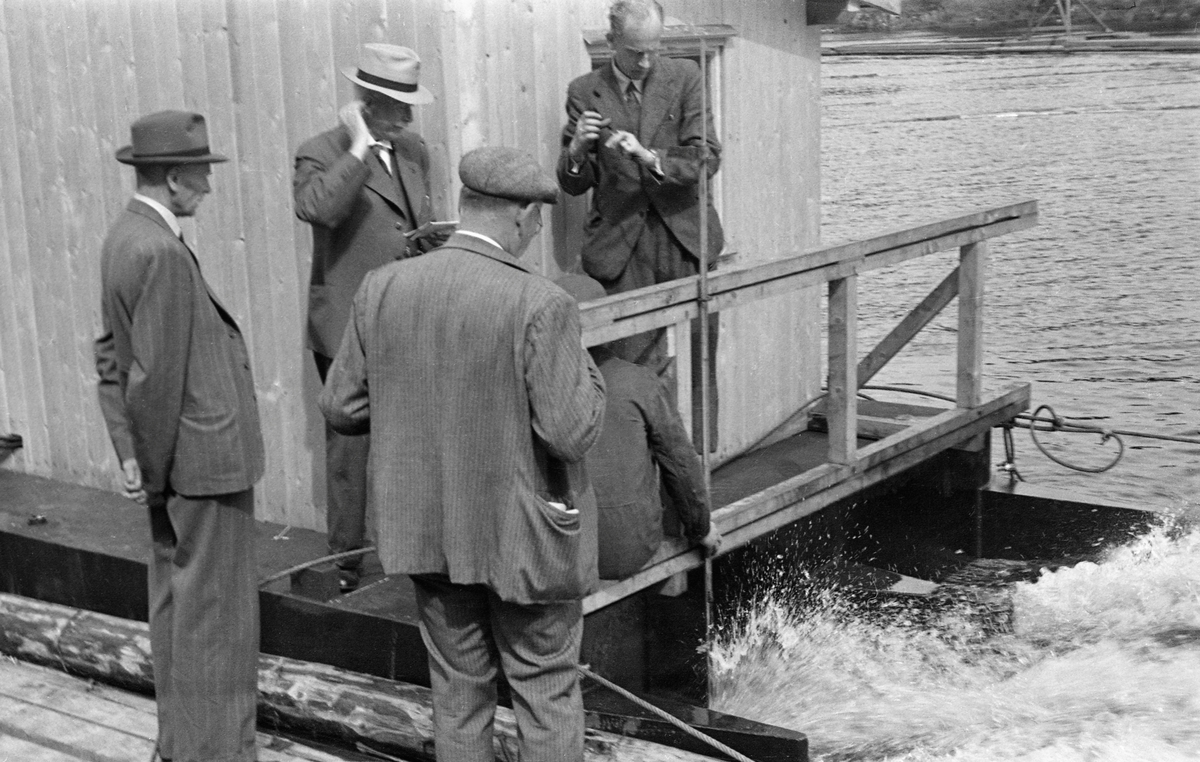 Fra forsøk med strømdannerapparat i dammen ved Kykkelsrud kraftstasjon i 1942. Fotografiet viser fem menn, hvorav en satt med ryggen mot fotografen og var delvis skjult, avbildet ved et lite hus som sto på pongtonger. Vannet mellom pontongene var kvitskummende og tydelig i bevegelse. Det som satte vannet i bevegelse var sannsynligvis en diger, roterende propell som befant seg i vannet, og som antakelig ble drevet av en motor som befant seg i det nevnte huset, via kjeder. Flåten ble holdt i posisjon ved hjelp av vaiere, som formodentlig var forankret i fastpunkter på land. Poenget var at propellen skulle skape en ønsket bevegelse i vannet. Her ved Kykkelsrud, hvor det var en kraftstasjon, dreide det seg antakelig om å skape strømninger som skulle bevege fløtingsvirket mot den tømmerrenna som skulle ta stokkene forbi kraftverksdammen. Det dreide seg med andre ord om et tiltak som skulle øke ekspedisjonshastigheten ved tømmerrenna uten å øke forbruket av vann, noe som jo ville ha gått på bekostning av kraftproduksjonen. Vi vet foreløpig lite om akkurat dette forsøket, men etter krigen markedsførte Olsen & Thorsen mek. verksted, ofte kortkortet til «OTHO», slike strømdannere med motorer som kunne yte fra 10 til 60 hestekrefter. Firmaet hevdet at disse maskinene kunne generere strøm så langt som 60 til 80 meter fra det stedet hvor den roterende propellen ble montert. Fløtingsfunksjonærene erfarte nok at vikrkningen ikke var fullt så vidtrekkende som produsenten forespeilte dem. Olsen & Thorsen mek. verksted markedsførte det samme produktet overfor virksomheter som hadde behov for mudring. 

Mennene på fotografiet - antakelig de fire dresskledde herrene - skal være (fra venstre): Kr. Mørch, W. Olsen, Myrvold og Mohn.