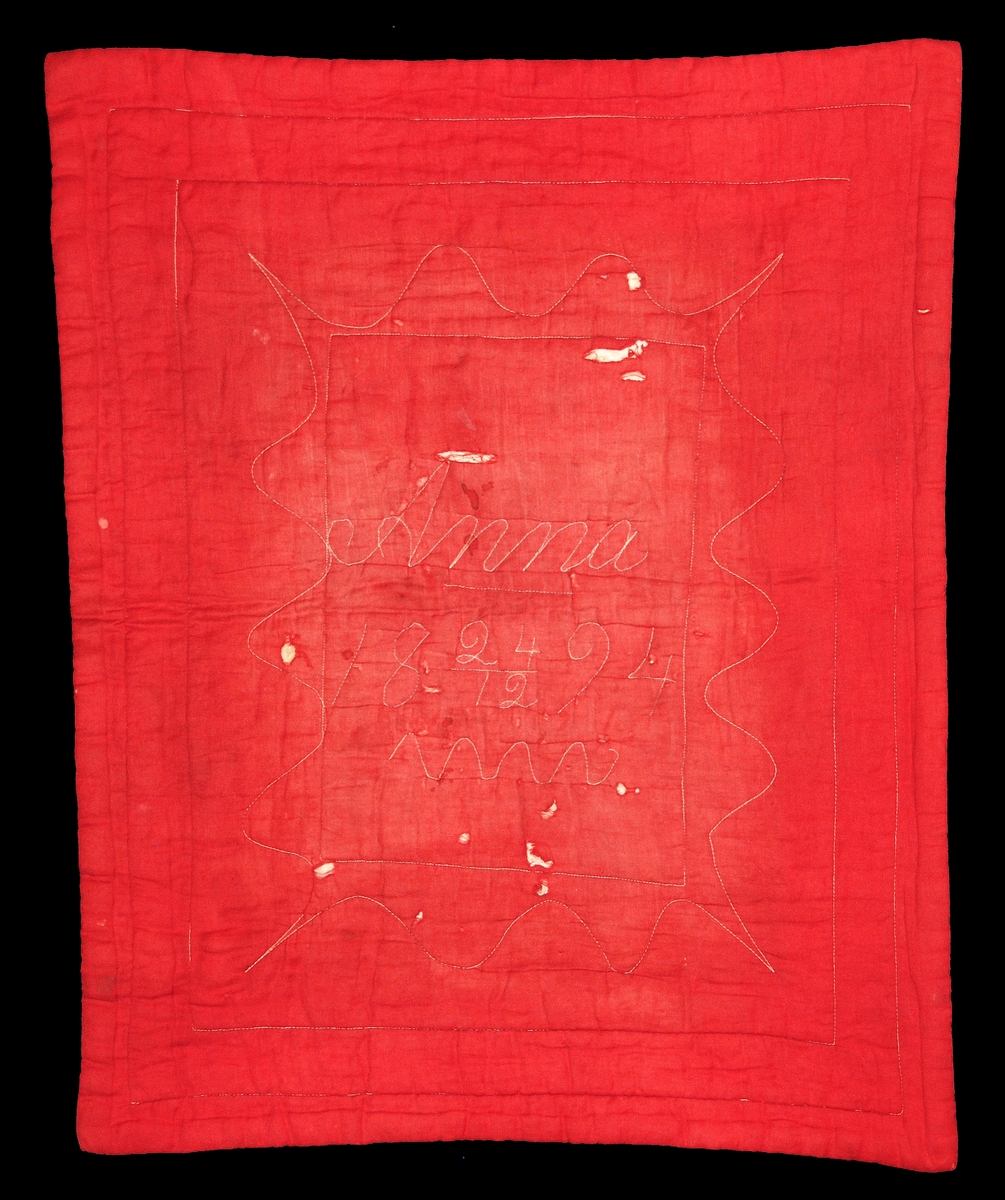 Täcke, för docksäng, rött bomullstyg. Stoppat med vadd. Märkt på ovansidan med sysmaskinsstygn "ANNA 24/12 1894". Undersidan av naturfärgat linne.