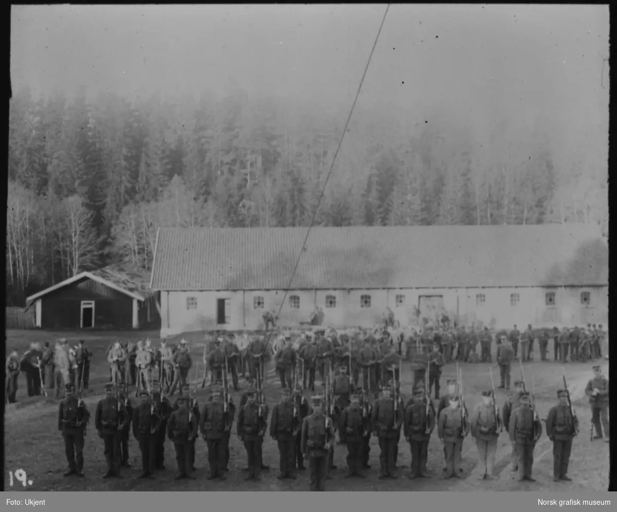 Soldater er stilt opp på et gårdstun, antageligvis i området mellom Kongsvinger og Skotterud. Det står flere soldater i bakgrunnen, men disse er ikke oppstilt.
