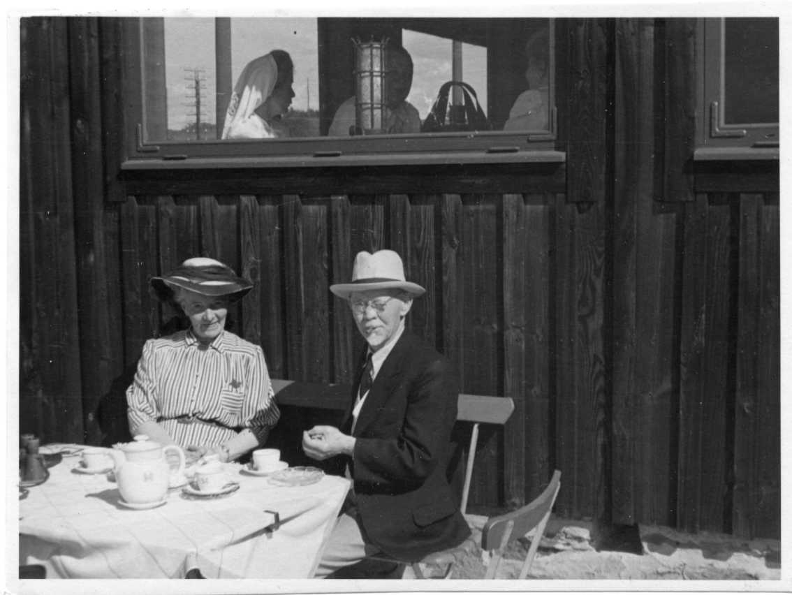 En äldre man och kvinna sitter i solsken vid ett kaffebord utmed en husvägg. Bägge har hatt, mannen en cigarill i munnen.