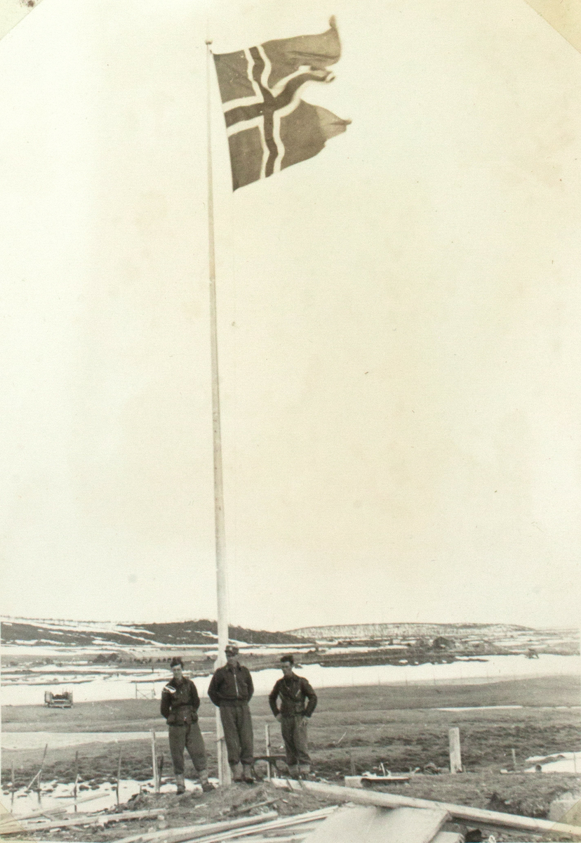 Tre menn står ved en høy flaggstang der norskeflagget veiver i vinden. Landskapet er delvis dekket med snø.