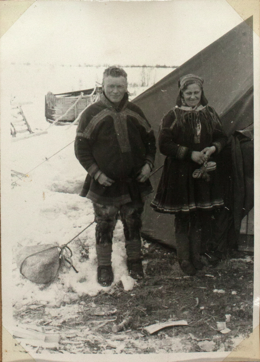 Bildet viser et ungt, samisk ektepar. De står ved siden av hverandre, foran et telt. De er ikledd tradisjonelle samiske antrekk. Teltet bak dem er festet med hjelp av stein. I bakgrunnen ser man en slede som ligger på side. Landskapet er snødekket.
