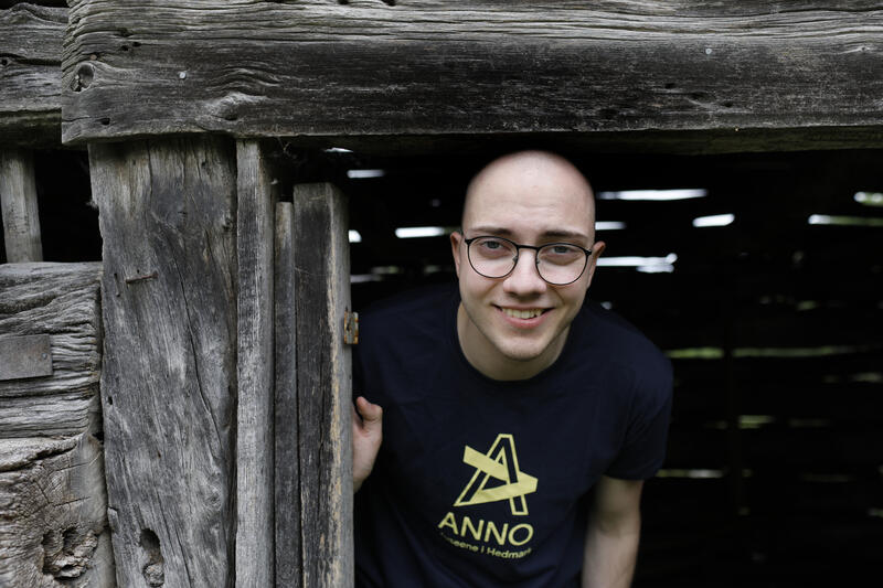 Skallet ung mann med Anno-trøye titter fram fra et grått tømmerhus.