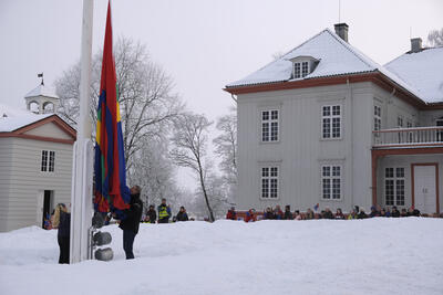 Vi ser fasaden til Eidsvollsbygningen, noen ti-talls mennesker foran huset følger med når to barn og en voksen heiser samisk flagg i hovedflaggstanga. Det er vinter og snø