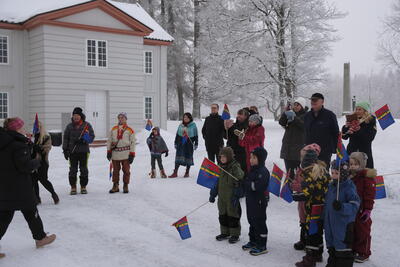 En forsamling mennesker i ulik aldre, noen i samekofte, andre i vinterklær. De står ved søndre sidebygning foran Eidsvollsbygningen. Det er vinter, de har samiske håndflagg.