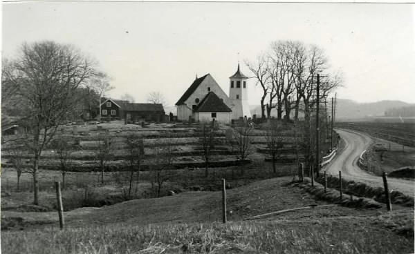 Älvsåker kyrka med klockstapel intill grusvägen som slingrar fram i landskapet. Kyrkogården är terrasserad.