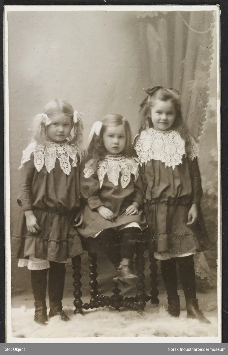 Visittkortfoto, tre barn fotografert i studio. Tre jenter, alle kledd i kjole med blondekrage rundt halsen. Alle har sløyfer i håret. Den yngste jenta sitter på stol i mellom de to andre jentene.