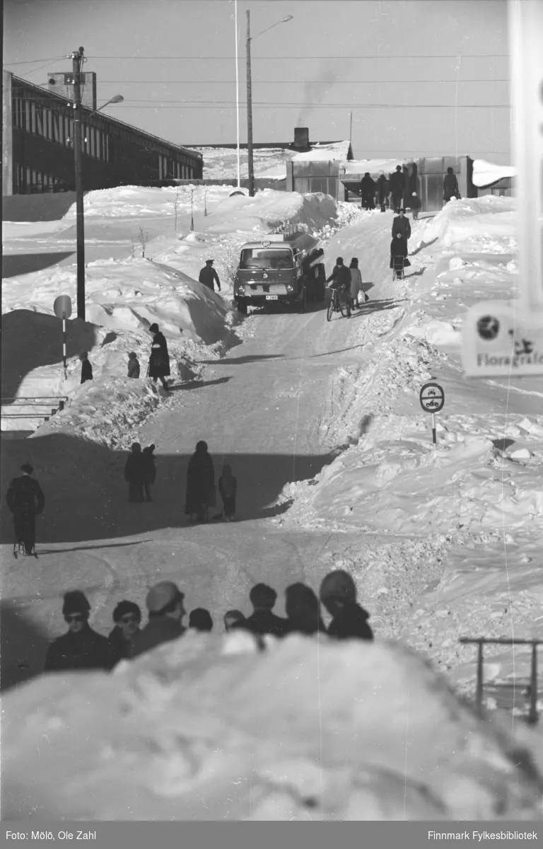 En vinterdag i Vadsø, ca. 1968. Folk og en bil parkert på bakken mot Statens hus (før innvandrermonumentet kom i 1977). Bilen er Bedford tankbil reg. på Y-2969. Leveranse av BP, muligens fyringsolje.
