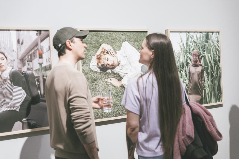 Two people stand in front of framed photographs on the wall. The photographs are portraits of people outdoors.