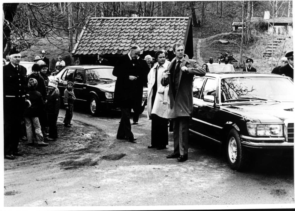 En Mercedes Benz följd av en SAAB parkerade i Röttle. Vid första bilen står kung Carl XVI Gustaf i ljus rock, till vänster länsråd Ragnar Forss. Poliser och allmänhet runtomkring.