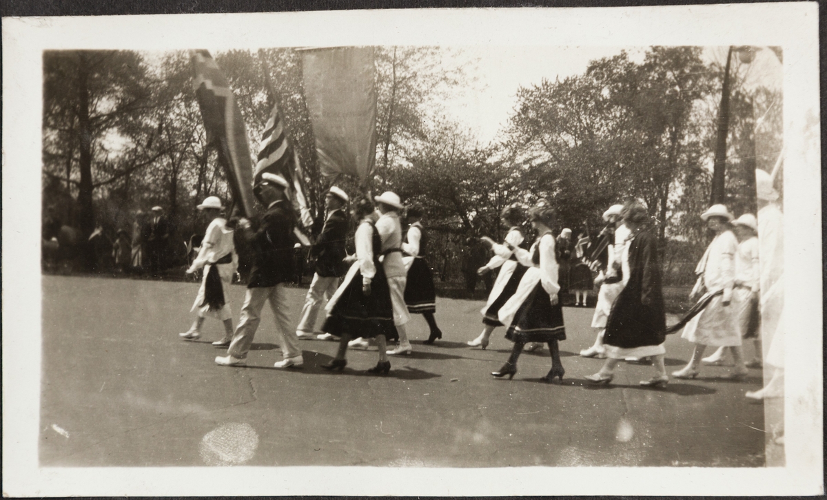 Folketog under syttende mai i USA. Kvinner i bunad og like drakter og menn i like dresser går foran med norsk og amerikansk flagg og ei fane.