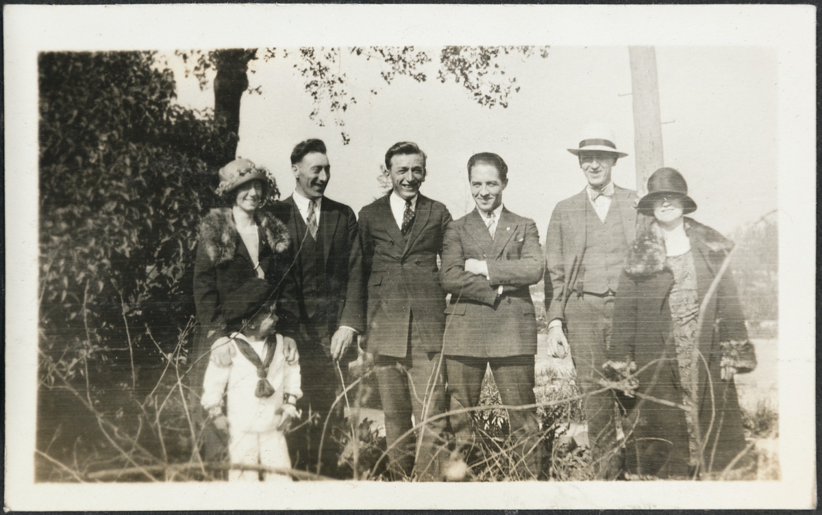 En gruppe menn, kvinner og et barn smiler til fotografen i en park. Fra venstre er mrs. Ragnhild Bang med sønnen Billy. TIl høyre for henne er Bjarne Fixdal, Kalle (Karl Bårdsen), Jakob Lura, mr. Bang og en ukjent kvinne.