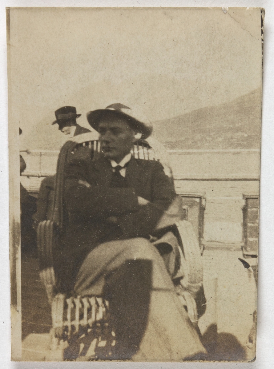 Portrett av mann sittende i stol på dekket av en båt. Mannen har hatt, dressjakke, hvit skjorte og slips. Andre passasjerer kan skimtes i bakgrunne. Man ser hav og land helt i bakgrunnen.