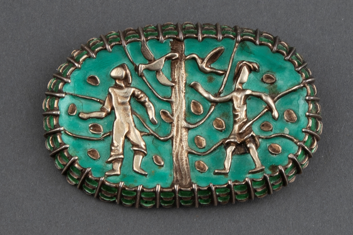 Oval brosje i sølv med turkis emalje. Motiv med to stiliserte menneskefigurer, to fugler og et tre med blader. Nål på baksiden.