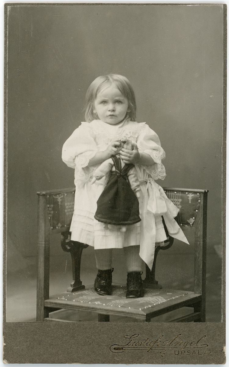 Kabinettsfotografi - liten flicka med en docka står på en stol, Uppsala