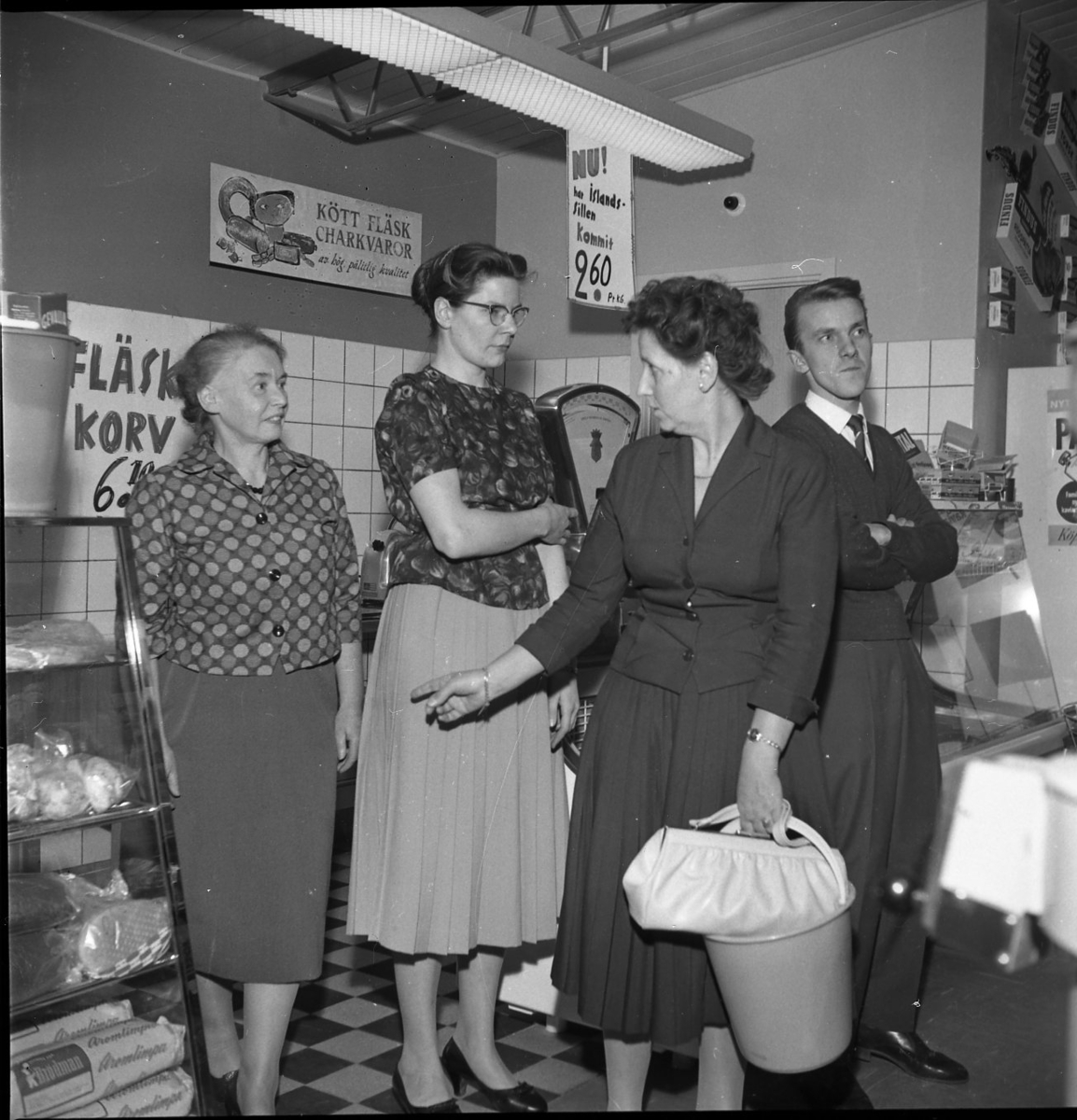 Tre kvinnor och en man står i en charkuteributik. De lyssnar till någon som står utanför bild. En kvinna håller en hink i handen. Damen till vänster kan heta Ryd i efternamn och arbetade i Skär- stads Handelsförening,
