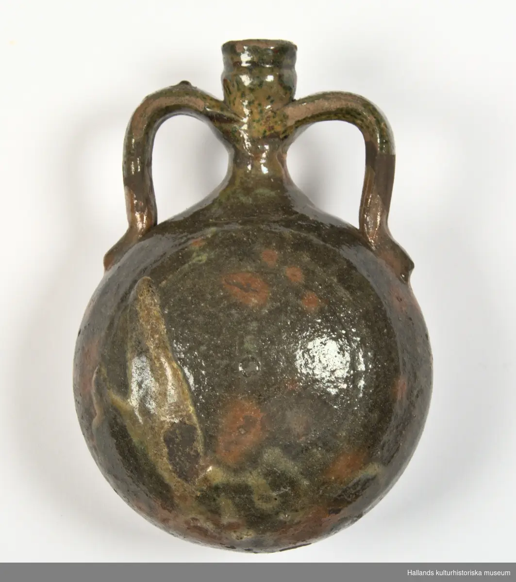 Flaska ("fältflaska") av keramik. Flaskan har formen av en fältflaska, plan på ena sidan och bukig på den andra. Med två öron, handtag, från halsen till livet. Grön- och gulglaserad.