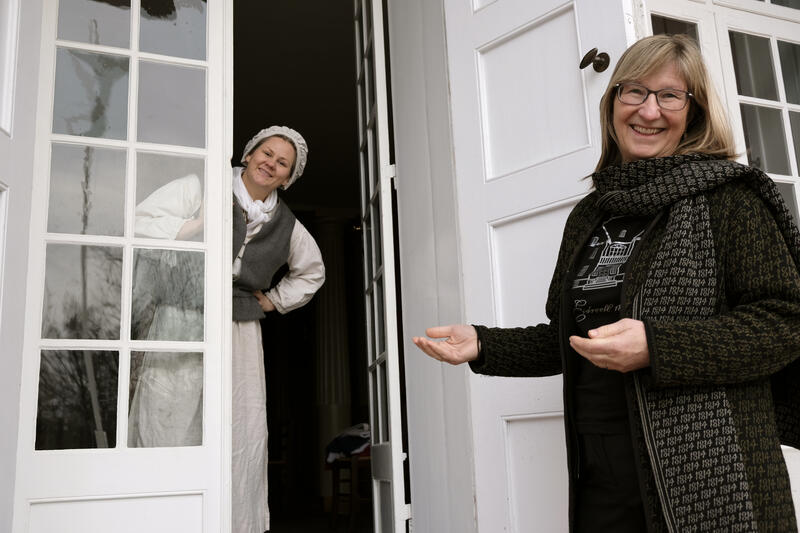 En kvinne i Eidsvoll 1814-klær ønsker velkommen, i bakgrunnen titter en kvinne ut av døra ikledd kostyme som illuderer at hun er en tjenestejente fra 1800-tallet.
