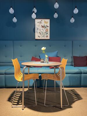Interiørbilde fra Kafe Standpunkt viser to stoler ved et bord. I bakgrunnen en stor, turkis sofa, på veggen en Grunnlovs-plakat og i taket henger flere små lamper