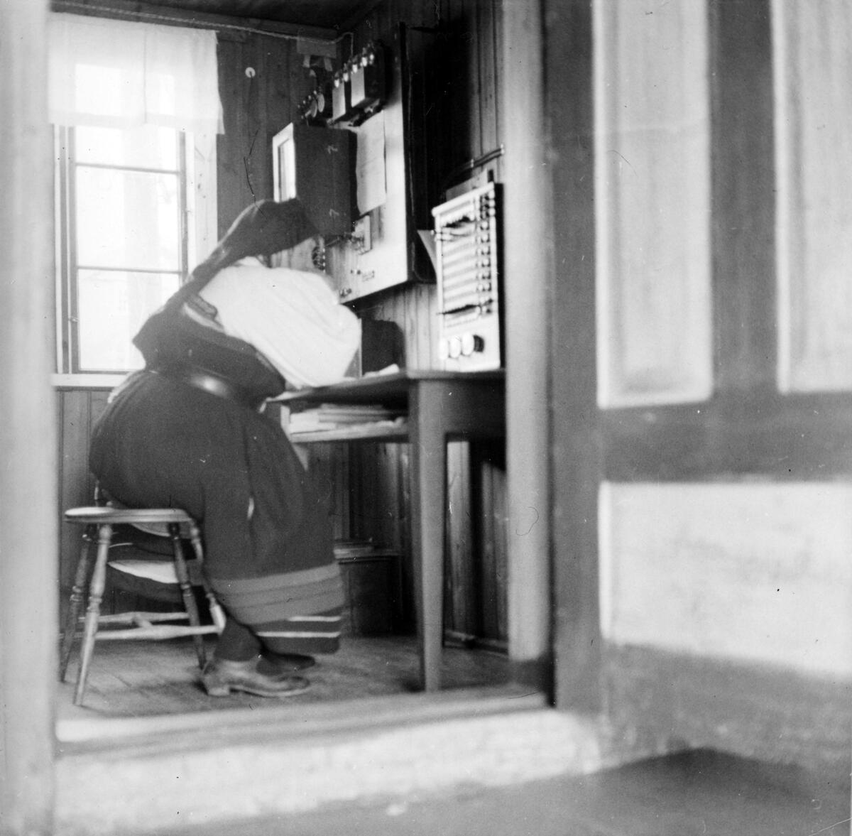 En kvinne i norsk folkedrakt sitter på en krakk i et lite rom, med telegrafutstyr. Svart-hvitt fotografi.