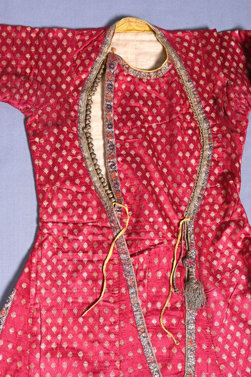 Mansrock/Kaftan från Rajasthan, Indien. Öppen mitt fram, slitsar i sidorna. 
Sydd av rött, diagonalmönstrat sidentyg med små broscherade blommor av guldfärgat lan. Kantad med två sorters mönstervävda band med snarlikt mönster med blommor och guldtråd - framtill, nedtill och i sidorna upp till slutet av sidslitsarna. Runt bröst- och halsöppning, ett flätat bland påsytt över det mönstervävda. Nederdelen fodrad med sidentyg, smalrandigt i gult och mörkblått, kanterna och kragen med enfärgat gult tyg. Bröstpartiet och ärmarna fodrade med ett glest beige bomullstyg samt det randiga tyget längst ned i ärmarna. Rocken är vadderad med bomullsvadd. Rundad urringning ned till midjan där rocken knyts ihop med smala gula band. Intill dessa, dekorativa snörmakeriarbetade tofsar fästa på vänster framstycke, varav den ena saknar själva tofsen. 31 stycken små dekorationsknappar av guldtråd längs bröstöppningen på höger sida. "Bröstlapp" av samma tyg som resten av rocken täcker "urringningen" och fästs uppe vid nacken med en liten rund knapp.