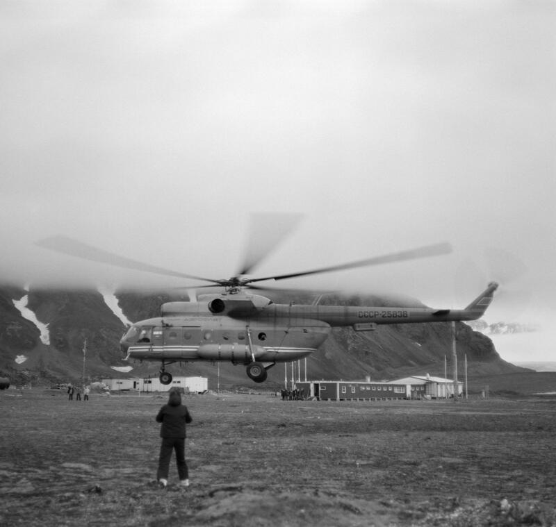 Bildet vise et sovjetisk helikopter som går inn for landing. En person står i forgrunnen med ryggen mot kamera. Fjell i bakgrunnen.