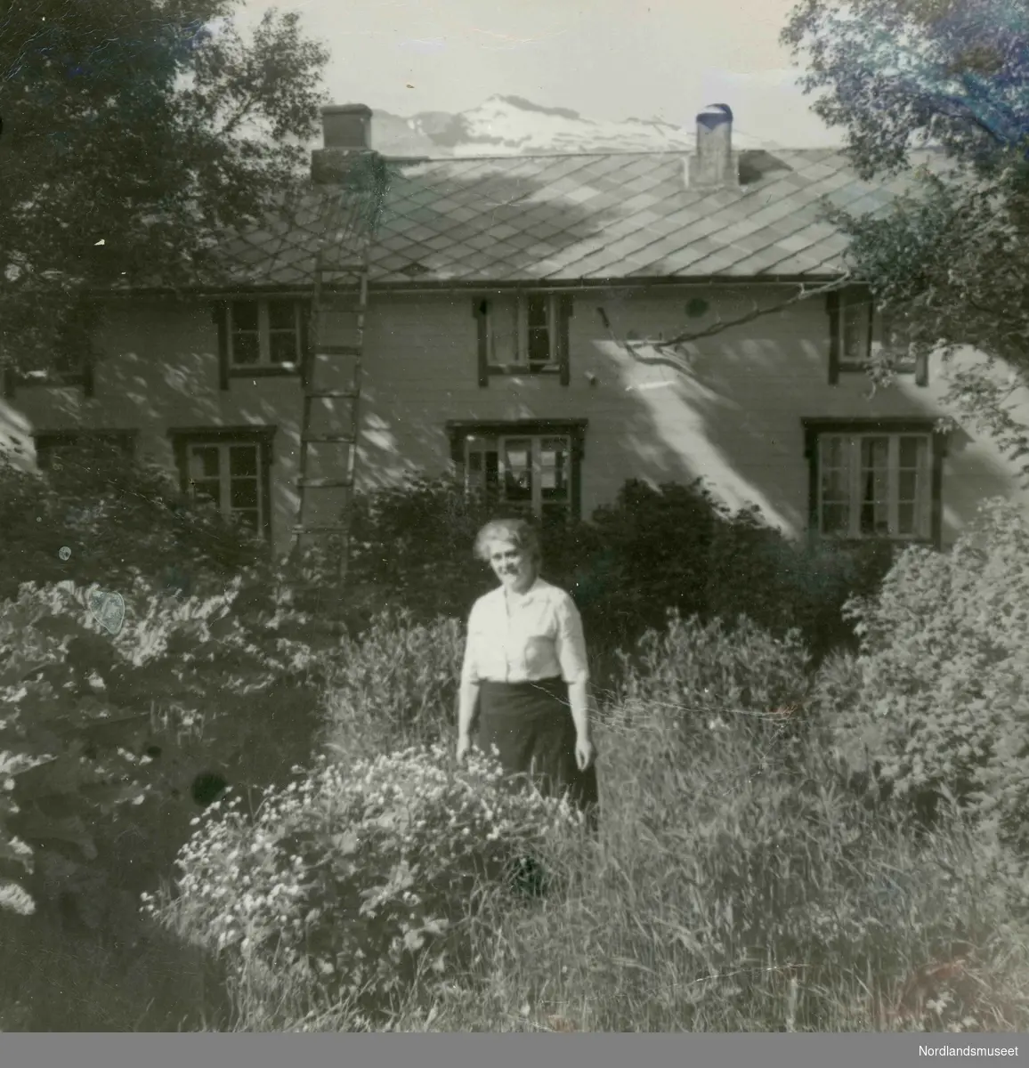 Eldre dame som står i hagen utenfor huset sitt. Gjertrud Larsen (f.1888) fra Myklebostad i Leiranger. Gift med William Kornelius Larsen. Huset inneholdt både posthus og telefonsentral. Over taket ses fjellet Småtindan.