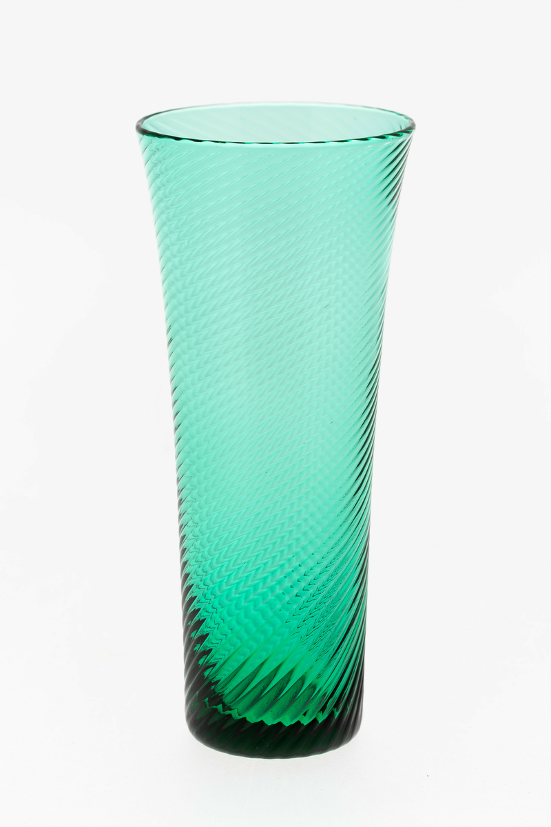 Seltersglass i grønnfarget gjennomskinnelig glass. Svak konisk form med sirkulær munningsrand, hvor spiralformede riller snor seg langs korpus.