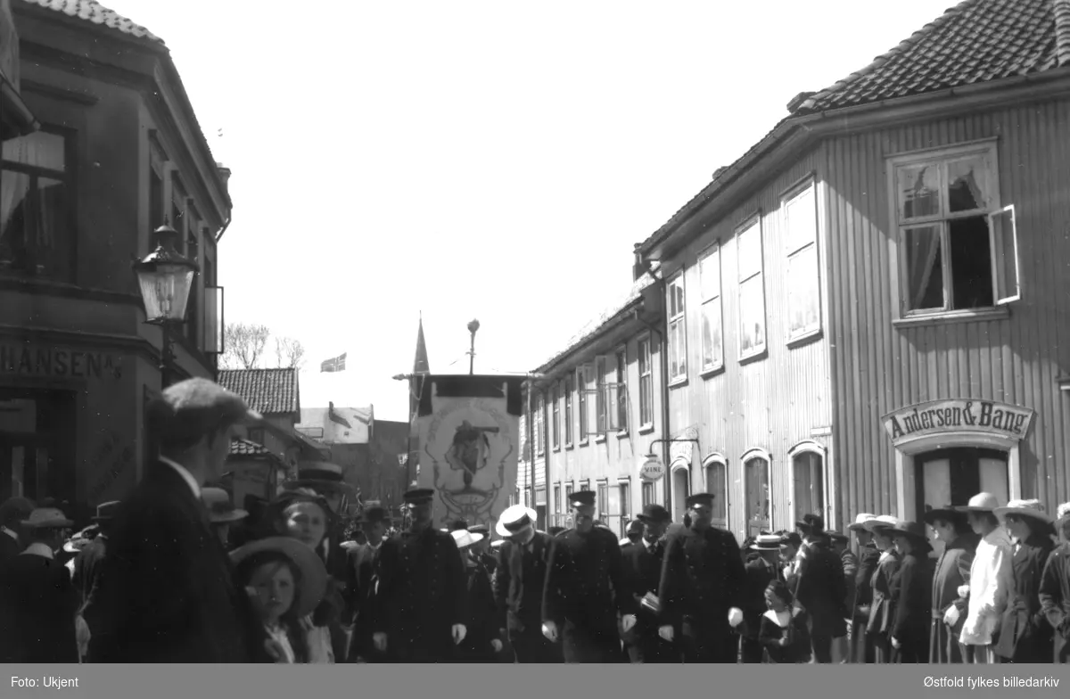 17. mai tog, fane med teksten: "Moss Høiere Almenskole", ca. 1920-25. Skilt: "Andersen & Bang","O. Hansen A/S, garn & Manufactur". Dronningensgate 1 på høyre side, hele høyre side er idag revet.