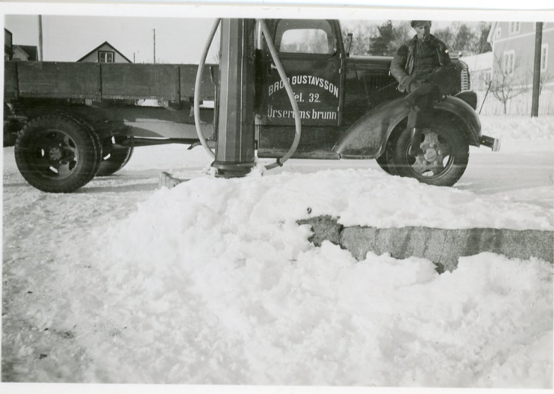 En man med pipa i munnen sitter på stänkskärmen på en lastbil med dörren märkt: "Bror Gustavsson Tel. 32. Örserums brunn". Den står vid Ingessons bensinpump i Örserum. Det är vinter med snö på marken.