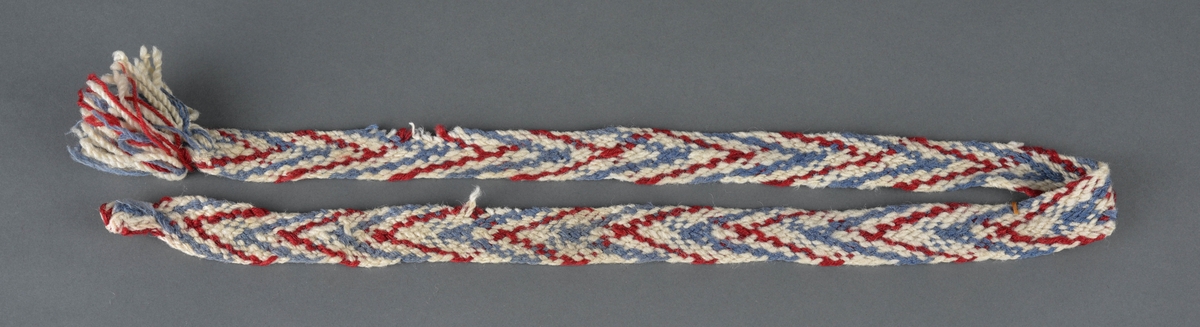 Strømpeband, hoseband fletta i rødt, blått og sauekvitt ullgarn.
Dusk i ene enden. Mangler den andre båndet.