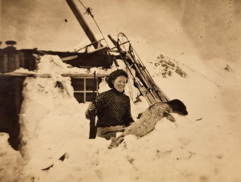 Bildet viser en smilende kvinne som holder en rifle. Hun står foran en enkel hytte, omgitt av store mengder snø.