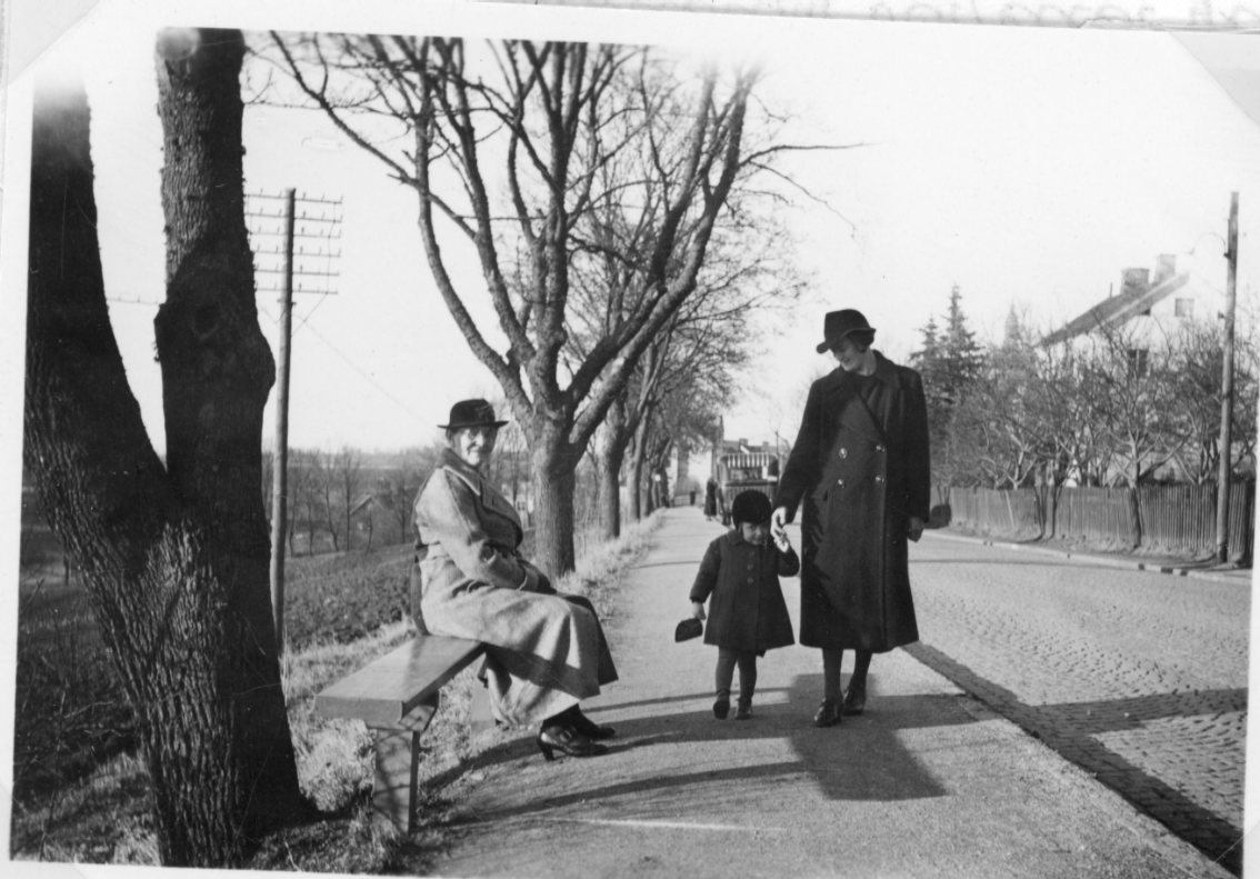 En äldre kvinna med hatt och kappa sitter på en bänk vid Grännas södra infart. En yngre kvinna med ett barn i handen står intill.