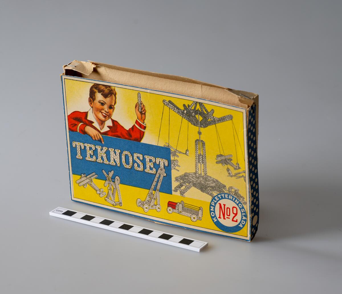 Originalförpackning för Teknoset, tom. Kompletteringslåda till byggsats. Framsida föreställande pojke med byggdel i handen samt byggexempel så som karusell, flygplan, turbin, stege samt bil med flak.