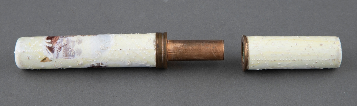 Sylinderformet nålehus i emalje og kobber, i to deler. Gulhvit med landskapsmotiv i flere farger.