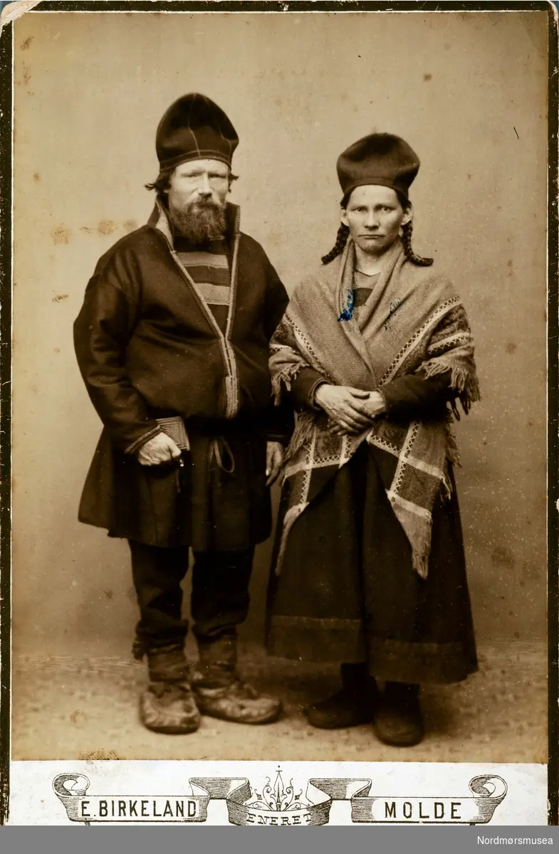Portrettfoto av et samisk par som kom til Romsdals amts byer for å selge sine reinsdyr i årene 1870 til 1880. Begge er kledt i hverdagsklær. Mannen ser ut til å holde en bibel eller en salmebok i hånden, mens hun står med et skjal over skuldrene. Selgeren kunne fortelle at ";de reiste rundt i mørebyene i 1880 årene, og stod på Skoleplana (nå Kongens plass) og solgte reinsdyrkjøtt. De kom hver vår, og ble godt kjent både med barn og voksne i byen. Min mor stod på bakerbutikk i Torvgata (Petersens bakeri) og fikk dette bildet av mannen på bildet ";Torkel"; og madamen. Mor var en venn av dette hyggelige ekteparet. Hun var datter av en Svendsen på Meløya."; Fotograf er E. Birkeland, Molde, i årene 1870 til 1880.