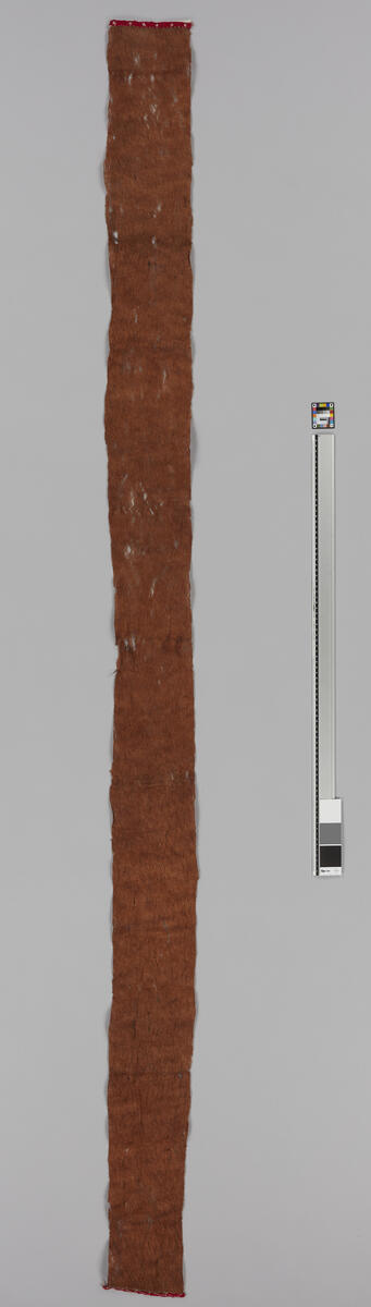 Fra Hovedkatalogen: Langt, smalt stykke brunt basttøy, på de korte sider kantet med smale røde tøistrimler. H: 237cm br: 16.0cm Borneo