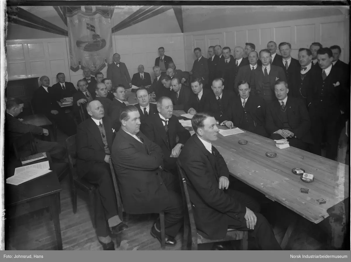 Gruppe menn rundt bord i annledning skøytetinget 1933. På veggen i lokalet hengen fanen til Snøgg Idrettsforening.