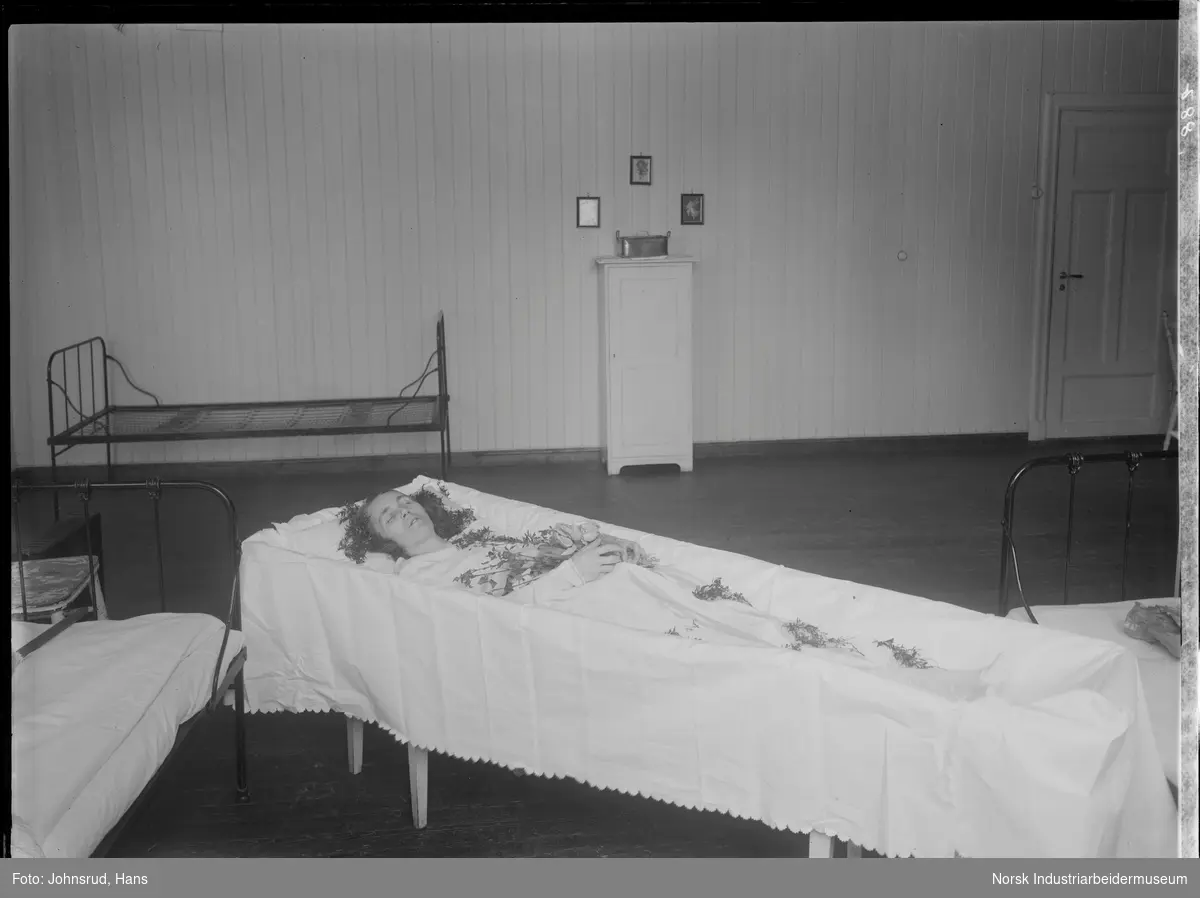 Post mortem fotografi av kvinne liggende i åpen kiste. Kvinnen er kledd i hvite klær og har blomster dekorert på og rundt seg. Kisten står i et rom med flere senger, muligens ett sykehusrom.